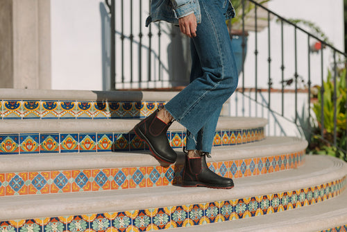 A person in rolled-up blue jeans and black boots walks up colorful, decorative tiled steps outdoors, with a black metal railing and greenery visible in the background.