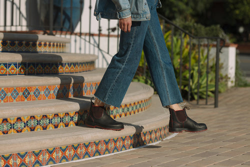 A person wearing blue jeans, a denim jacket, and black boots with red elastic sides walks down colorful tiled steps outdoors. Only the lower half of their body is visible.