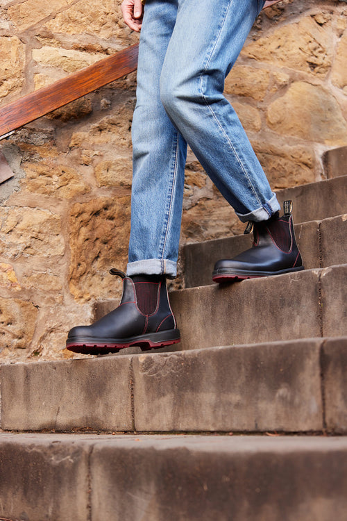 Person wearing cuffed blue jeans and black Chelsea boots with red soles, standing on outdoor stone steps next to a stone wall with a wooden railing.