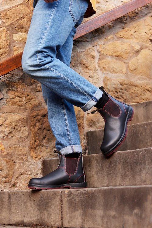 Person wearing rolled-up blue jeans and dark brown Chelsea boots with red stitching, standing on outdoor concrete stairs beside a stone wall. Only the lower legs and feet are visible.