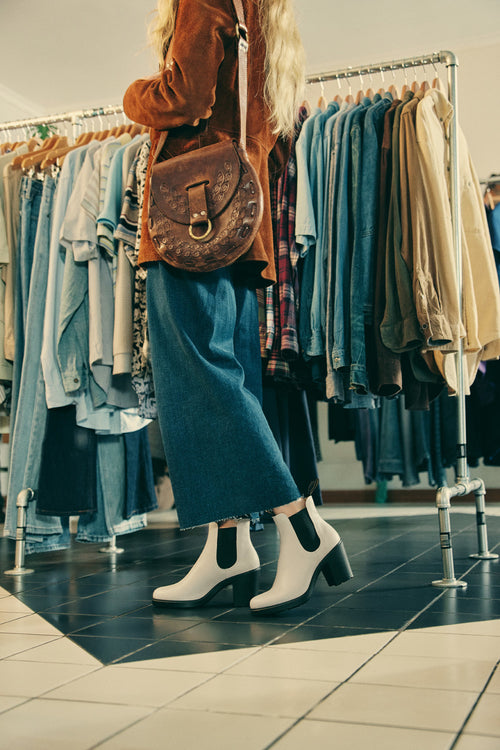 A person with long blonde hair, wearing a brown jacket, blue jeans, white ankle boots, and a brown shoulder bag, stands in front of racks of assorted clothing in a store.