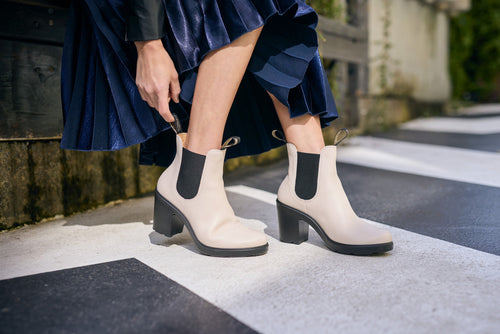 A person in a blue pleated skirt adjusts light-colored heeled ankle boots with black elastic side panels while standing on a crosswalk.