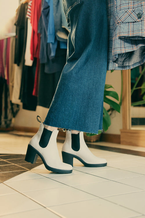 Person wearing wide-leg blue jeans and cream-colored heeled Chelsea boots stands indoors on a tiled floor, holding a plaid shirt. Clothing racks and a plant are visible in the background.