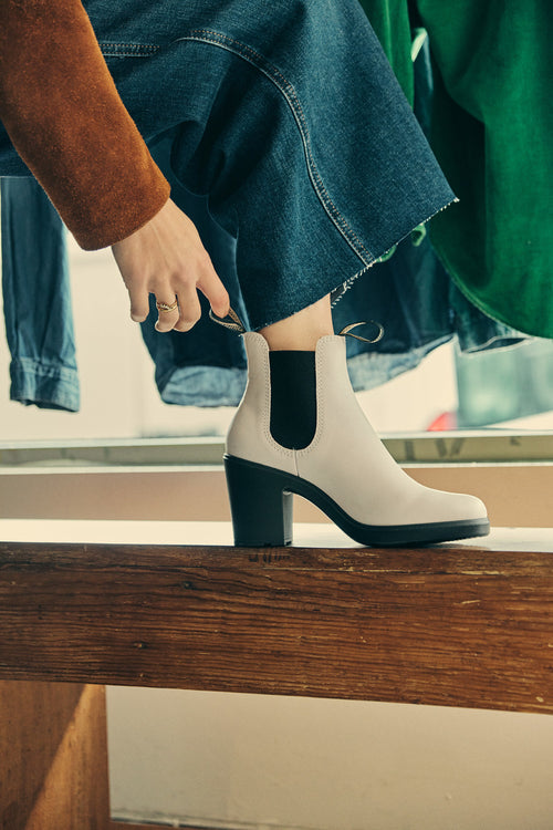 A person wearing a brown jacket and denim skirt puts on a white heeled ankle boot with black elastic panels, sitting on a wooden bench near a window.