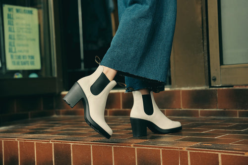A person wearing cropped blue jeans and beige heeled ankle boots with black elastic sides is walking up brown tiled steps outside a building.