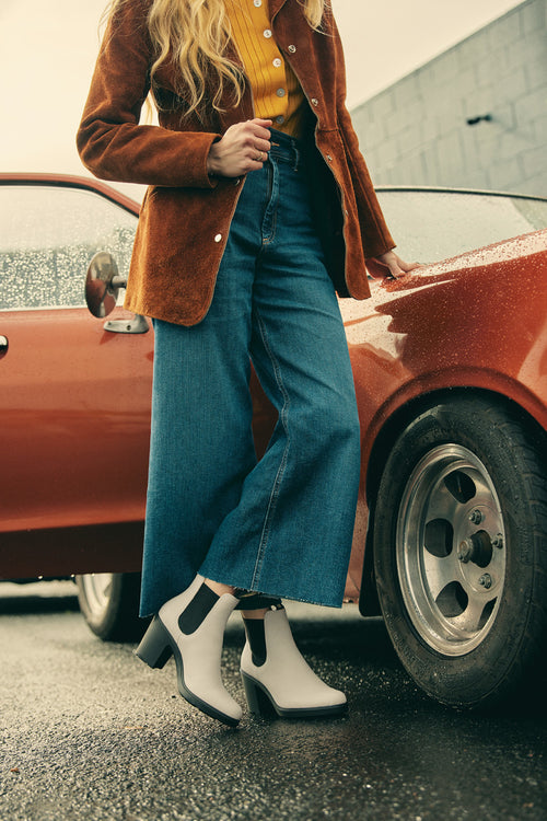 A person wearing a brown jacket, mustard shirt, wide-leg jeans, and white ankle boots stands beside a vintage red car on a wet pavement. The scene has a retro, stylish vibe.