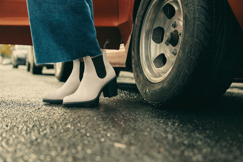 A person wearing white heeled ankle boots and blue jeans stands next to the wheel of a red car on a wet, paved street.