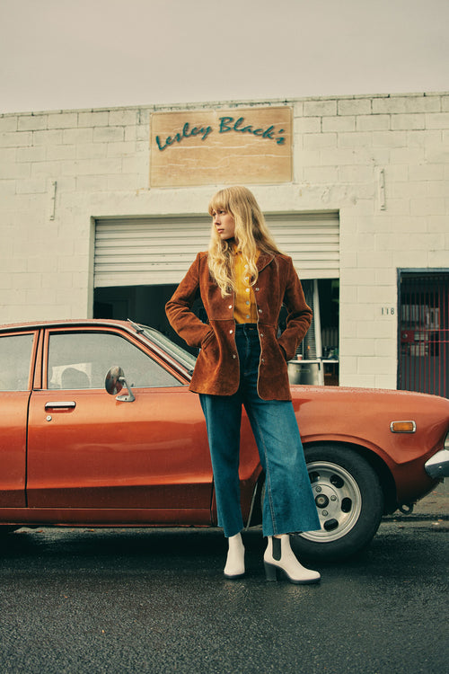 A woman with long blonde hair, wearing a brown jacket, yellow shirt, wide-leg jeans, and white boots, stands in front of a vintage orange car parked outside a white brick building with a wooden sign.
