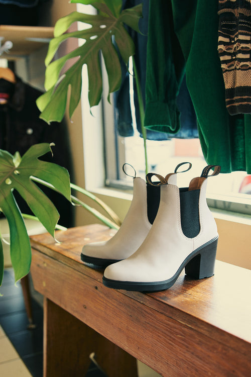 A pair of white heeled ankle boots with black elastic sides sit on a wooden bench beside green leafy plants and hanging clothing in a stylish indoor setting.