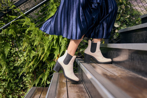 A person wearing a pleated navy skirt and stylish cream ankle boots with black elastic sides is walking up outdoor stairs, with green ferns and plants visible along the railing.