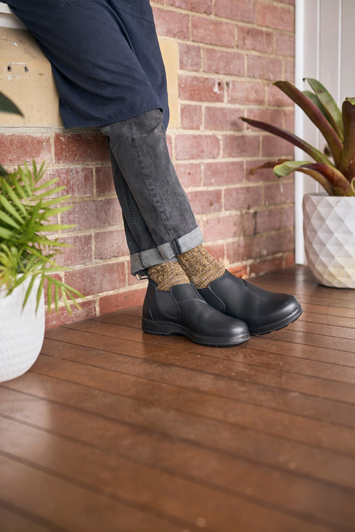 Person in dark pants and rolled cuffs sits on a ledge, showing off black ankle boots and brown patterned socks. Wooden floor, brick wall, and potted plants are in the background.