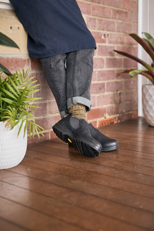 A person wearing rolled-up dark jeans, patterned socks, and black slip-on work boots stands with legs crossed on a wooden porch next to potted plants, with a brick wall in the background.