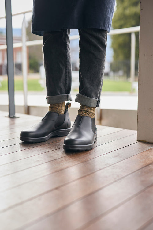 A person wearing rolled-up dark jeans, brown patterned socks, and black slip-on leather boots stands on a wooden floor near a railing with a grassy area visible outside.