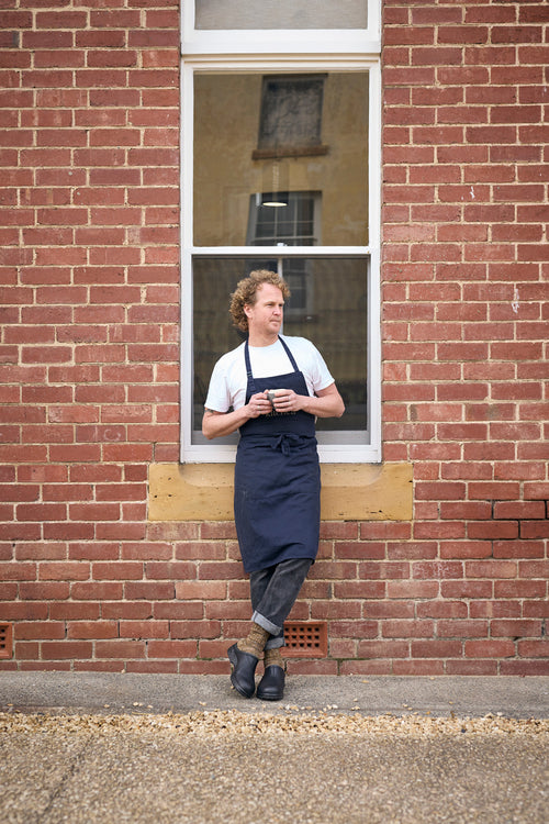A man wearing a white shirt, dark apron, and jeans stands casually against a brick wall beneath a window, holding a cup and looking to the side.