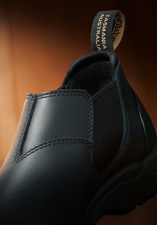 Close-up of a black slip-on shoe with visible stitching and a pull tab at the back that reads TASMANIA AUSTRALIA in yellow text. The background is warmly lit in brown tones.