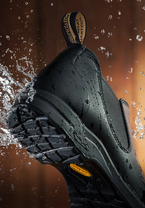 Close-up of a black waterproof shoe with visible tread, water splashing off it, and a branded pull tab, highlighting its durability and water resistance.
