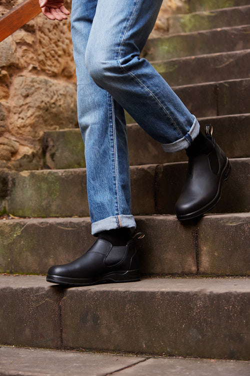 Person wearing rolled-up blue jeans and black ankle boots is walking up outdoor stone steps next to a rough stone wall. Only the lower half of the body is visible.