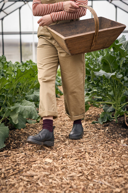 A person wearing tan pants and boots walks through a garden greenhouse, holding a wooden basket by their side. Green leafy plants grow on both sides of a woodchip-covered path.