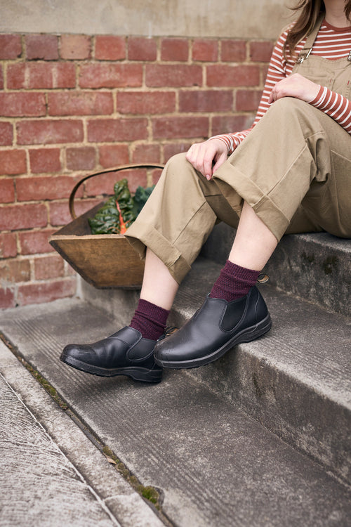 A person wearing beige overalls, a striped shirt, maroon socks, and black slip-on shoes sits on outdoor steps next to a metal basket filled with leafy greens, with a brick wall in the background.