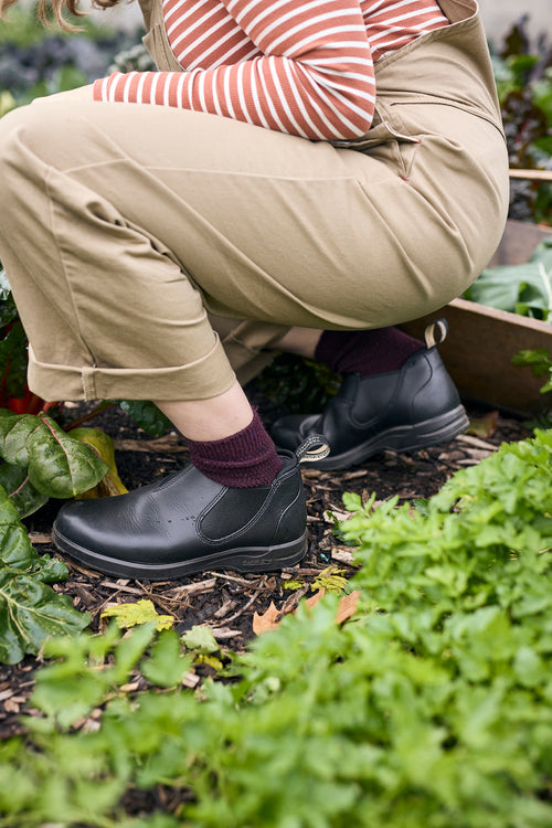 A person in khaki pants, a striped shirt, and black shoes squats among green leafy plants in a garden, tending to the plants.