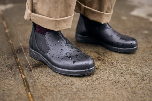 Close-up of a person wearing black waterproof boots and beige pants standing on a wet sidewalk, with water droplets visible on the boots.