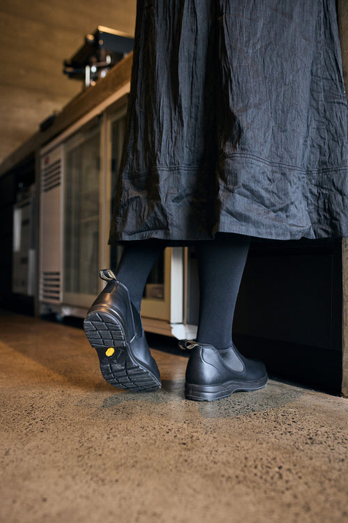 A person wearing a long black skirt, black tights, and black slip-on shoes stands in a kitchen with a polished concrete floor, facing a counter.