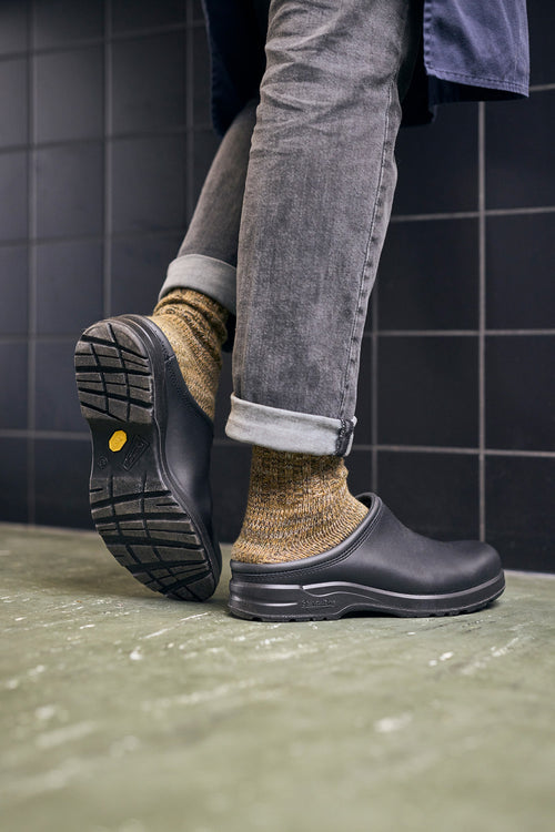A person wearing gray rolled-up jeans, yellowish wool socks, and black slip-on clogs stands on a greenish floor with one foot lifted, showing the sole. The background features black tiled walls.