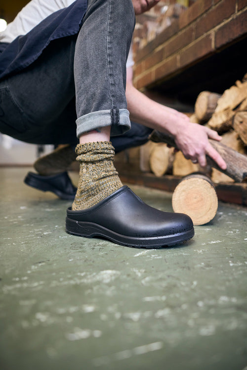A person wearing gray jeans, patterned socks, and black clogs crouches by a stack of firewood, reaching to pick up a log. The background shows a brick wall and more firewood.