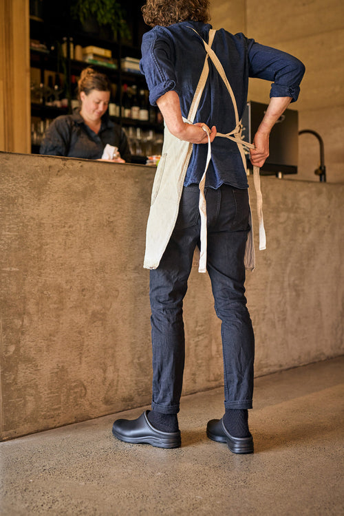 A person in dark clothing and an apron ties the apron behind their back, standing in a modern cafe. Another person is at the counter in the background, partially visible.