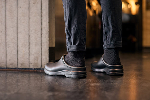 A person wearing black slip-on shoes and dark socks stands indoors on a shiny floor, with the legs of their dark pants slightly rolled up. Only their lower legs and feet are visible.