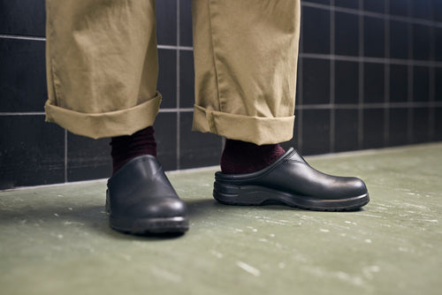 A person wearing rolled-up khaki pants, dark socks, and black slip-on clogs stands on a green floor in front of a black tiled wall.