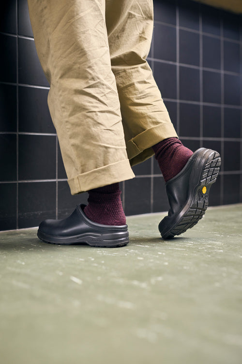 Person wearing beige pants, maroon socks, and black slip-on clogs stands on a green floor with one foot lifted, against a black tiled wall.