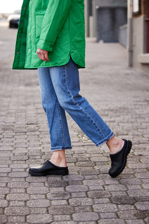 Person walking on a cobblestone sidewalk, wearing a green quilted coat, blue jeans, and black slip-on shoes. Only the lower half of their body is visible.