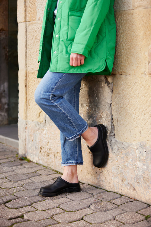 A person wearing a bright green quilted jacket, cuffed blue jeans, and black slip-on shoes leans against a stone wall with one foot resting on the wall and the other on the cobblestone ground.