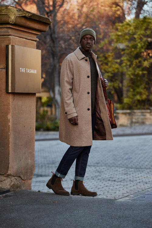 A man wearing a beige coat, dark jeans, brown boots, and a green beanie stands on a sidewalk near a sign that reads The Tasman, holding a brown bag over his shoulder. Trees with autumn leaves are in the background.