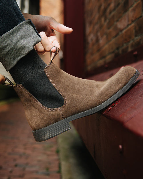 A person wearing dark socks and brown suede Chelsea boots adjusts their pant leg, standing on a red metal ledge next to a brick wall.