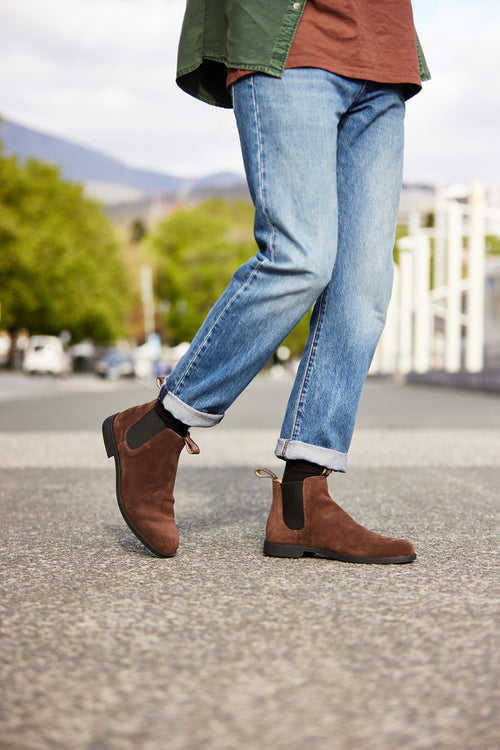 A person wearing rolled-up blue jeans, a brown shirt, a green jacket, and brown suede Chelsea boots is walking outdoors on a paved surface, with trees and mountains in the background.