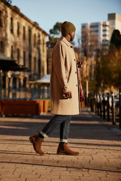 A man wearing a beige coat, brown beanie, jeans, and brown boots walks along a sunlit city street with buildings and greenery in the background.
