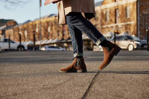 A person wearing rolled-up jeans, a beige coat, and brown suede ankle boots is walking on a paved street with blurred buildings and parked cars in the background.