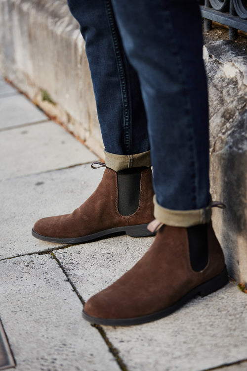 A person wearing dark rolled-up jeans and brown suede Chelsea boots stands on a stone sidewalk near a stone wall.