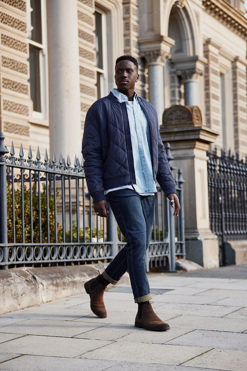 A man walks on a city sidewalk near an ornate building, wearing a navy quilted jacket, light blue shirt, cuffed jeans, and brown suede boots. He appears confident and relaxed.