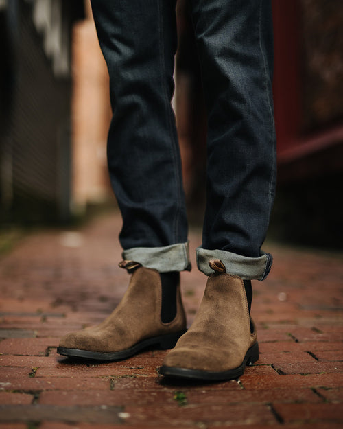 Person wearing cuffed dark jeans and brown suede Chelsea boots, standing on a wet brick pathway in an outdoor urban setting. The photo is focused on the lower legs and feet.