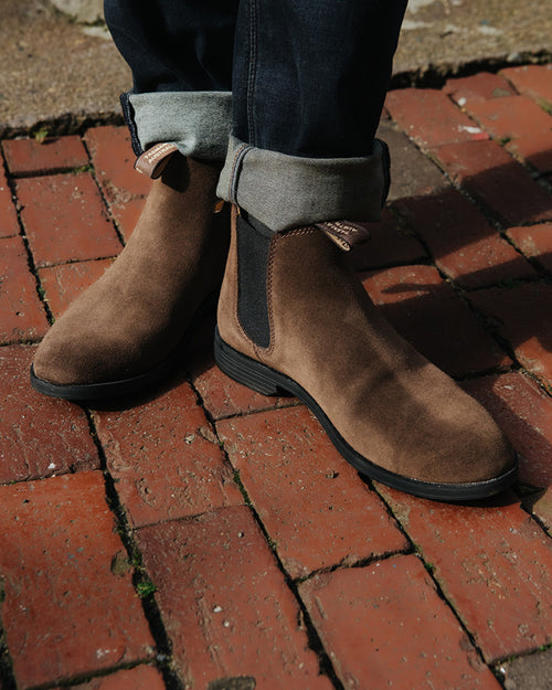 A person wearing cuffed jeans and brown suede Chelsea boots stands on a red brick pavement in sunlight.