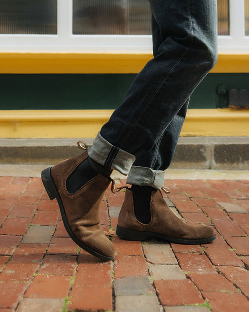 A person wearing dark rolled-up jeans and brown suede Chelsea boots walks on a red brick sidewalk in front of a yellow-and-green painted building.
