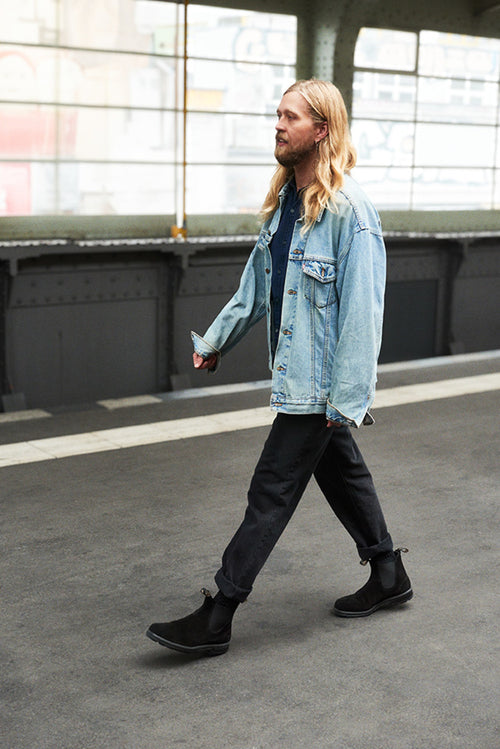 A man with long blond hair walks on a train platform. He is wearing a blue denim jacket, dark pants, and black boots. The background is slightly blurred, showing windows and graffiti.
