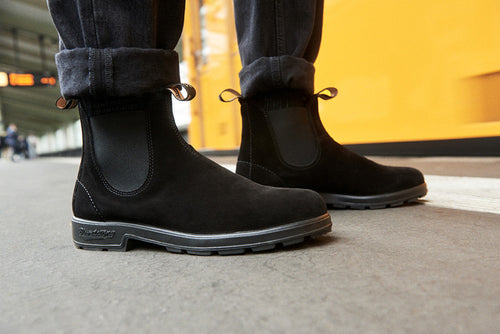 A person wearing black suede Chelsea boots and dark rolled-up jeans stands on a concrete surface, with a bright yellow wall in the background.