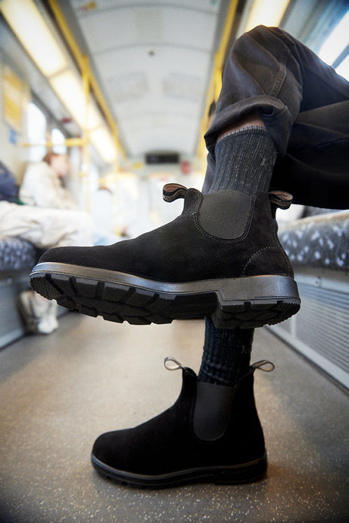 A close-up of a person wearing black Chelsea boots with thick soles, dark socks, and brown pants, sitting on public transport with their legs crossed. The train interior is slightly blurred in the background.