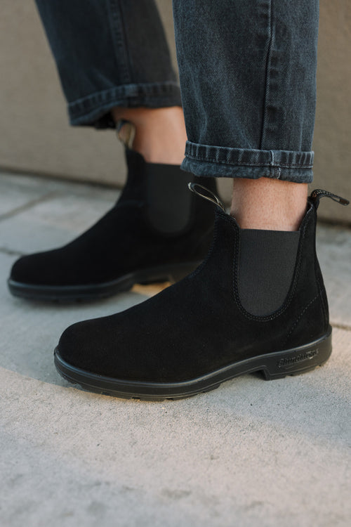 Person wearing black suede Chelsea boots with elastic side panels and black jeans with rolled-up cuffs, standing on a light-colored pavement.