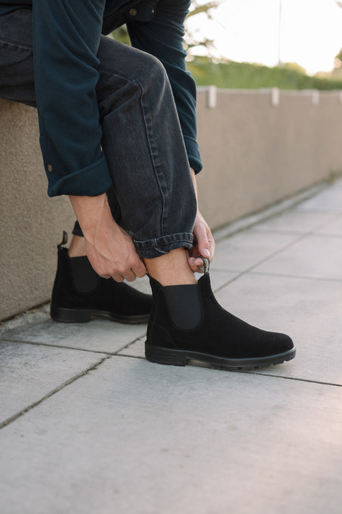Person sitting on a ledge outdoors, wearing dark jeans and a blue shirt, pulling up a black Chelsea boot on their foot. The scene is set on a tiled sidewalk with greenery in the background.