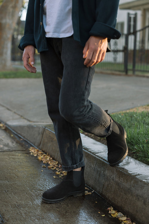 A person wearing black boots, dark jeans, a white shirt, and a dark jacket stands on a wet curb beside a grassy area and a sidewalk, with one foot slightly raised.